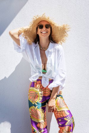 Photo for Portrait shot of an attractive woman wearing straw hat and sunglasses while standing at white wall. Laughing woman enjoying sunshine. - Royalty Free Image