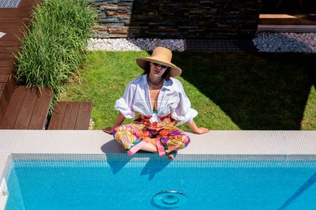 Foto de Happy woman wearing white shirt and straw hat while relaxing by the poolside. Full length shot. Copy space. - Imagen libre de derechos