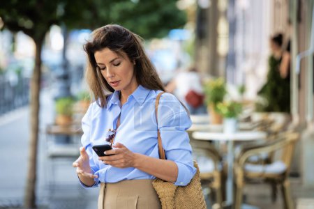 Photo for Brunette haired pretty woman standing the street and using mobile phone. Attractive female wearing business casual and text messaging. - Royalty Free Image