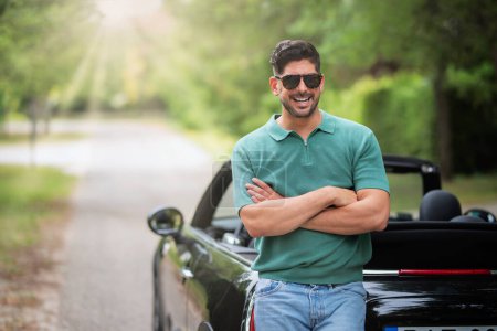 Foto de Un apuesto hombre de mediana edad de pie junto a su coche descapotable y riendo. Hombre seguro con gafas de sol y ropa casual - Imagen libre de derechos