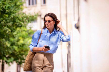 Foto de Mujer de cabello castaño con ropa casual de pie en la calle y mensajes de texto. Atractiva hembra con gafas de sol y sonrisa alegre. - Imagen libre de derechos