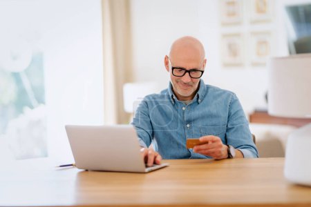 Téléchargez les photos : Homme d'âge moyen assis à table et ordinateur portable et payant par carte de crédit. Homme confiant portant des vêtements et des lunettes décontractés. - en image libre de droit