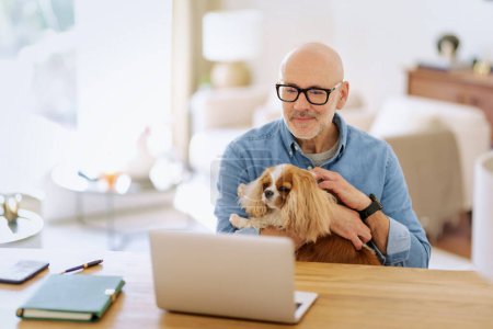 Foto de Un hombre de mediana edad sentado en una mesa con su cachorro y teniendo videoconferencia. Hombre seguro que usa ropa y gafas casuales. Oficina en casa. - Imagen libre de derechos