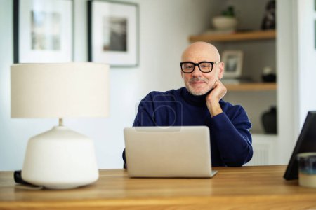 Téléchargez les photos : Homme d'âge moyen assis à table et utilisant un ordinateur portable pour le travail ou ayant un appel vidéo. Homme confiant portant des vêtements et des lunettes décontractés. Bureau à domicile. - en image libre de droit