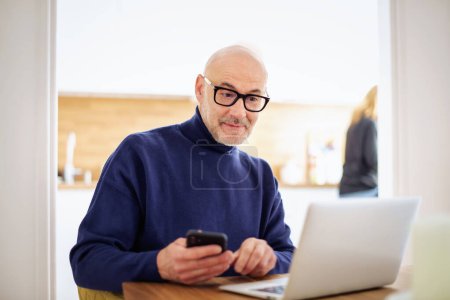 Photo for Thinking man text messaging and using laptop while working from home. Confident male sitting in the kitchen and text messaging. Home office. - Royalty Free Image