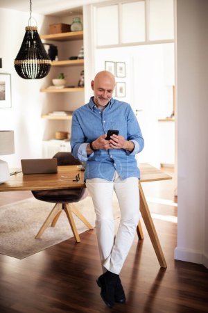 Photo for Happy middle aged man standing in the living room at home and using smartphone . Confident male wearing casual clothes and text messaging. Full length shot. Home office. - Royalty Free Image