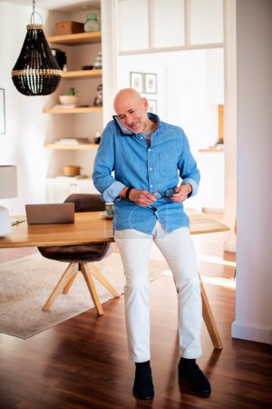 Photo for Happy middle aged man standing in the living room at home and making a call. Confident male wearing blue shirt and white jeans. Home office. Full length shot. - Royalty Free Image