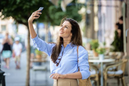 Photo for Brunette haired woman with smartphone standing on the street and taking a selfie with her phone or having video call. Attractive female cheerful smiling and wearing casual clothes. - Royalty Free Image