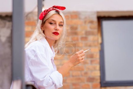 Foto de Primer plano de la hermosa mujer joven que usa bufanda para el cabello y camisa blanca mientras fuma cigarrillos y está de pie en el balcón. - Imagen libre de derechos