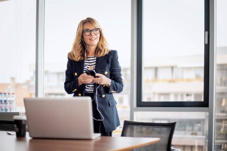 Photo for Mid aged businesswoman standing in a modern office and having video call. Confident professional woman using earphone and smartphone and having video call. - Royalty Free Image