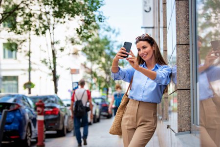 Foto de Mujer morena con el teléfono inteligente de pie en la calle y tomar una selfie con su teléfono o tener videollamada. Atractiva mujer alegre sonriente y con ropa casual. - Imagen libre de derechos