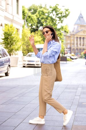 Photo for Attractive woman raising hand to call a taxi while walking on the street and having a phone call. Brunette haired female wearing shirt and sunglasses. Full length shot. - Royalty Free Image