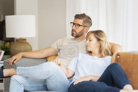 Foto de Una pareja de mediana edad está sentada en el sofá de su casa. Hombre guapo está viendo la televisión y la rubia está durmiendo a su lado.. - Imagen libre de derechos