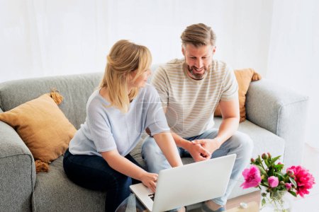 Photo for Middle-aged couple sitting at home on the sofa and using a laptop. Blond haired woman and grey haired man surfing the internet or having video call. - Royalty Free Image