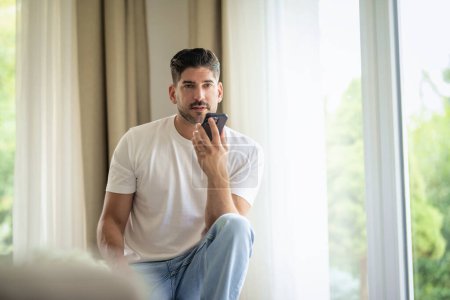Photo for Handsome man sitting on the couch at home and using his cellphone. Confident male wearing casual clothes and having a call. - Royalty Free Image