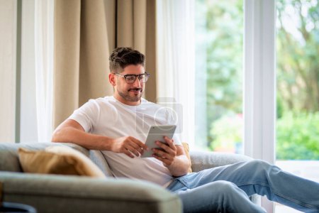 Photo for Middle-aged man sitting at home on the sofa holding a digital tablet in his hand. Confident male wearing glasses and casual clothes. He is having video call or browsing on the internet. Home office. - Royalty Free Image