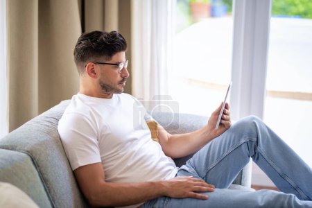 Foto de Hombre de mediana edad sentado en casa en el sofá sosteniendo una tableta digital en su mano. Hombre seguro con gafas y ropa casual. Está teniendo videollamadas o navegando por Internet. Oficina en casa. - Imagen libre de derechos