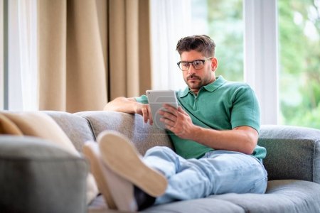 Photo for Middle-aged man sitting at home on the sofa holding a digital tablet in his hand. Confident male wearing glasses and casual clothes. He is having video call or browsing on the internet. Full length shot. - Royalty Free Image