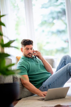 Photo for Handsome man sitting at home on the floor and using laptop. Confident male wearing casual clothes and looking at camera. - Royalty Free Image