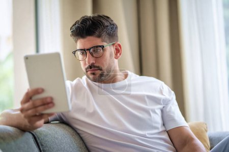 Photo for Close-up of a middle-aged man sitting at home on the sofa and holding a digital tablet in his hand. Confident male wearing glasses and casual clothes. He is having video call or browsing on the internet. Home office. - Royalty Free Image