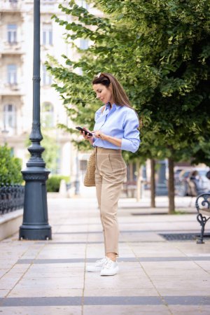 Photo for Brunette haired pretty woman walking down the street and using mobile phone. Attractive female wearing business casual and text messaging. Full length shot. - Royalty Free Image
