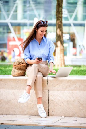 Photo for Brunette haired woman sitting on a bench in the city and using a laptop and smartphone for work. Happy mid aged female wearing sunglasses as casual clothes. Full length shot. - Royalty Free Image