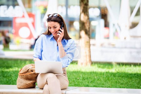 Photo for Brunette haired woman sitting outdoor in the city and using a laptop and smartphone for work. Confidner female having a call. Happy mid aged female wearing sunglasses as casual clothes. - Royalty Free Image