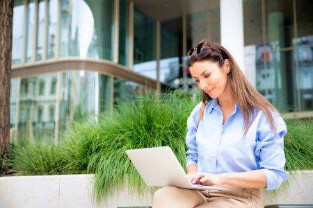 Photo for Brunette haired woman sitting on a bench in the city and using a laptop for work. Happy mid aged female wearing sunglasses as casual clothes. - Royalty Free Image