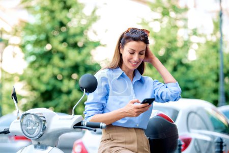 Foto de Mujer riendo con su scooter de pie en la calle de la ciudad y mensajes de texto en el teléfono móvil. Morena de pelo femenino vistiendo camisa azul. - Imagen libre de derechos