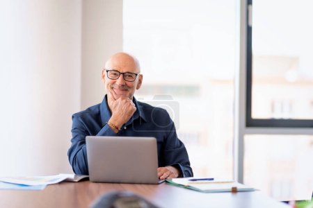 Téléchargez les photos : Homme d'affaires d'âge moyen assis à table dans un bureau moderne et utilisant un ordinateur portable pour le travail. Homme professionnel portant des lunettes et une chemise. - en image libre de droit