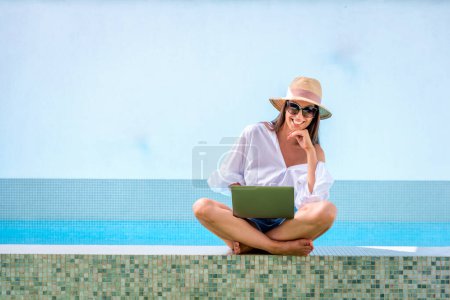 Foto de Querida mujer sonriente con sombrero de sol y camisa blanca y el uso de la computadora portátil en la piscina. tiro de longitud completa. Copiar espacio. - Imagen libre de derechos