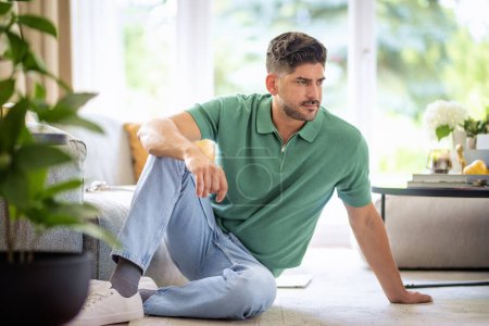 Téléchargez les photos : Toute la longueur d'un homme d'âge moyen assis sur le sol à la maison. Homme pratique portant des vêtements décontractés et se relaxant dans le salon. - en image libre de droit