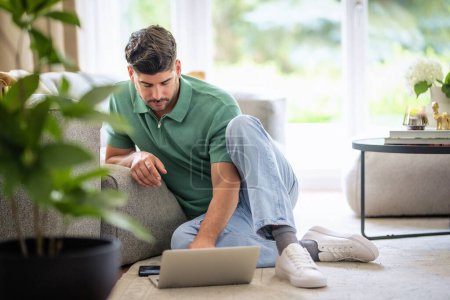 Photo for Mid aged man sitting on the floor in the living room at home and using laptop for work. Home office. Full length shot. - Royalty Free Image