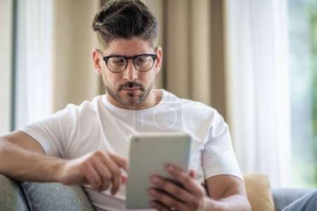 Téléchargez les photos : Gros plan d'un homme avec sa tablette numérique assis sur le canapé à la maison et au travail. Bureau à domicile. - en image libre de droit