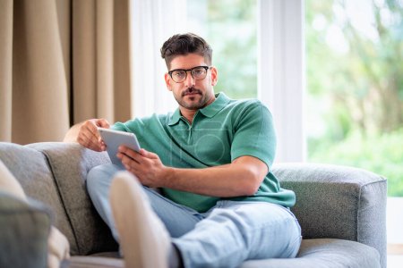 Téléchargez les photos : Un homme avec sa tablette numérique assis sur le canapé à la maison et au travail. Bureau à domicile. Plan complet. - en image libre de droit