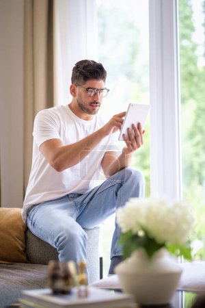 Téléchargez les photos : Un homme avec sa tablette numérique assis sur le canapé à la maison et travaillant ou ayant un appel vidéo. Bureau à domicile. Plan complet. - en image libre de droit