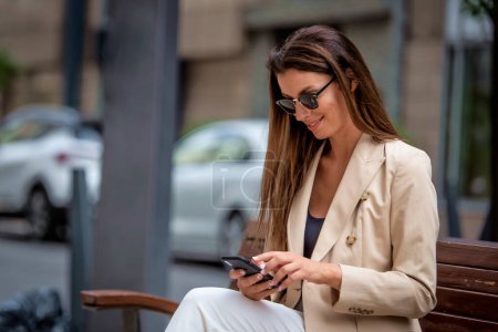 Photo for Brunette haired woman sitting on bench in the city and using mobile phone. Attractive female wearing business casual and text messaging. - Royalty Free Image