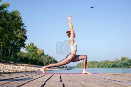 Foto de Longitud completa de mujer caucásica practicando yoga en el muelle junto al río. Atractiva hembra tiene el pelo rubio y sonriente. - Imagen libre de derechos