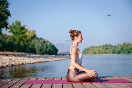 Téléchargez les photos : Toute la longueur de la femme caucasienne pratiquant le yoga sur la jetée près de la rivière. Attrayant femelle a les cheveux blonds et souriant. - en image libre de droit