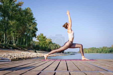 Téléchargez les photos : Toute la longueur de la femme caucasienne pratiquant le yoga sur la jetée près de la rivière. Attrayant femelle a les cheveux blonds et souriant. - en image libre de droit