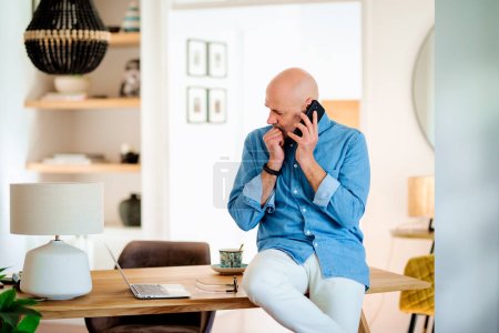Photo for Confident middle aged man making a call and using laptop while working from home. Confident male sitting in the dining room and having a call. Home office. - Royalty Free Image