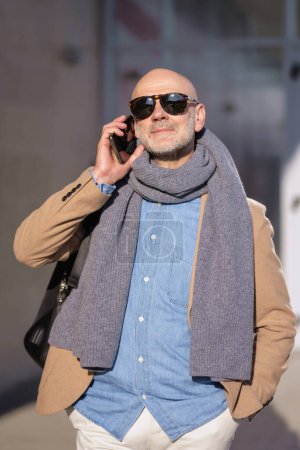Foto de Mature businessman using mobile phone in the city. Man in casual clothes walking outdoors by a building. - Imagen libre de derechos