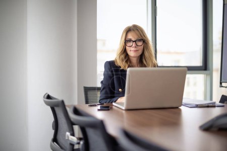 Photo for Businesswoman with laptop sitting at desk in a modern office. Confident professional woman working on laptop or having web conference. - Royalty Free Image