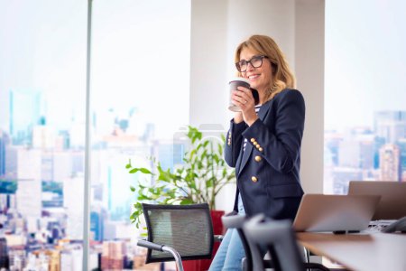 Photo for Portrait of mid adult woman standing by the window at the office and having coffee break. Confident female wearing blazer and glasses. - Royalty Free Image