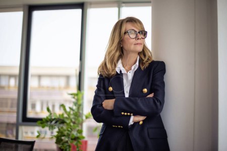 Foto de Mujer de negocios segura de pie con los brazos cruzados en una oficina moderna. Mujer profesional que usa gafas y chaqueta. - Imagen libre de derechos