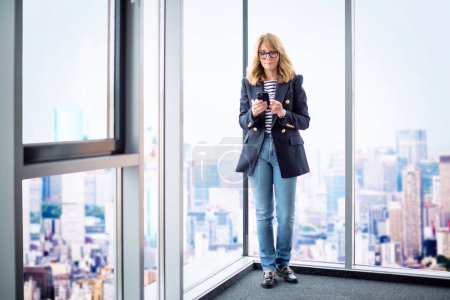 Photo for A middle-aged businesswoman standing in the office with the view of the city and using smartphone. Confident professional woman wearing blazer and text messaging. Full length shot. - Royalty Free Image