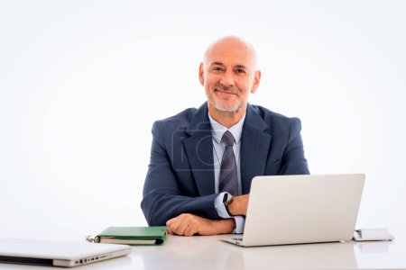 Téléchargez les photos : Homme d'affaires d'âge moyen assis au bureau et utilisant un ordinateur portable pour travailler dans un contexte isolé. Homme professionnel confiant portant costume et cravate. Espace de copie. - en image libre de droit