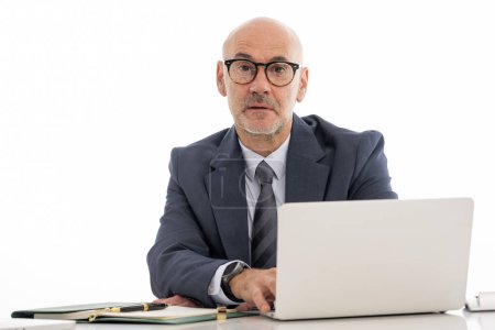 Photo for Mid aged businessman sitting at desk and using notebook for work against isolated background. Confident professional man wearing suit and tie. Copy space. - Royalty Free Image