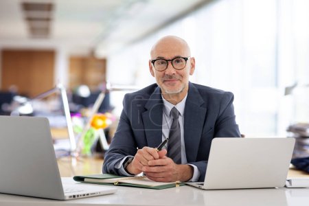 Téléchargez les photos : Homme d'affaires d'âge moyen assis dans le bureau et utilisant des carnets de notes pour le travail. Homme professionnel confiant portant costume et cravate. - en image libre de droit