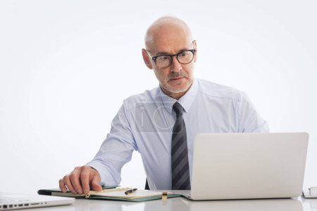 Téléchargez les photos : Homme d'affaires d'âge moyen assis au bureau et utilisant un ordinateur portable pour travailler sur fond blanc isolé. Homme professionnel confiant portant chemise et cravate. Espace de copie. - en image libre de droit